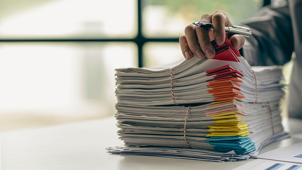 Closeup image of a large stack of papers organized by color. A person's hand holds a pen and flips through the papers.