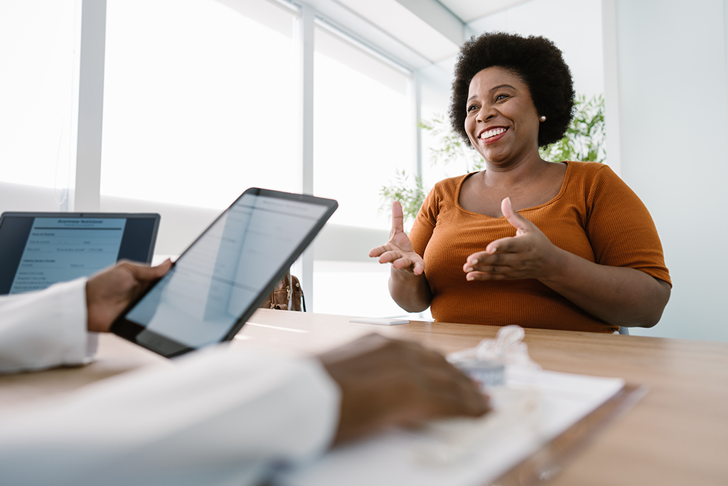 A middle aged African American woman smiles from across a table. She is gesturing to an African American doctor whose arms and hands are seen holding a tablet and paperwork.