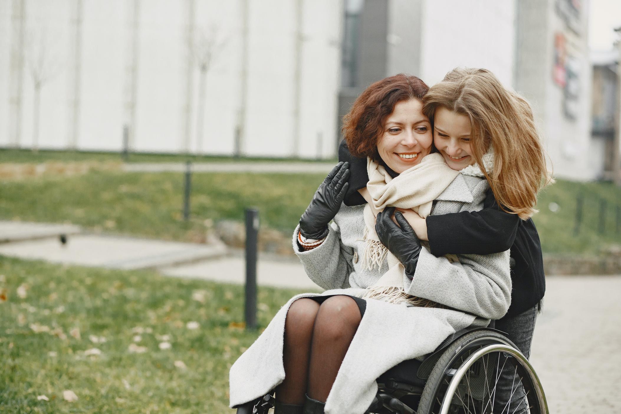 Woman in long winter coat, scarf and gloves sitting and smiling in a wheelchair as a young girl smiles and hugs her from behind.