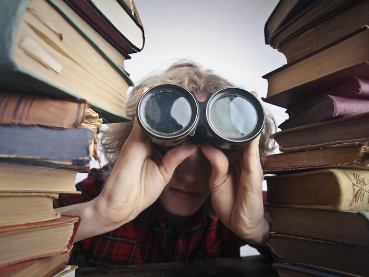 A person looking through binoculars between two towering stacks of old books.