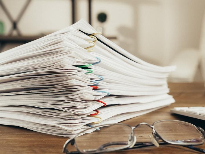 Stacked papers grouped together with multicolored paperclips on a wooden desk next to glasses and a wireless keyboard.