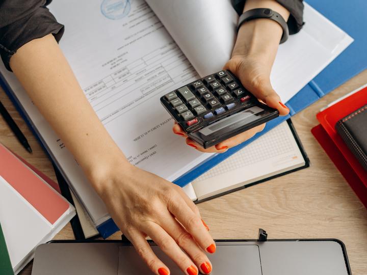 Person in black shirt at desk looking at paper files, calculator and laptop.