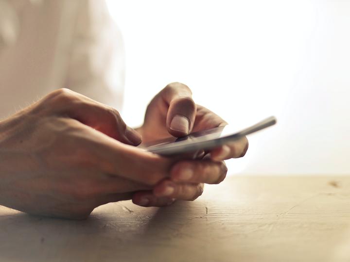 Person holding cell phone with both hands on table in front of bright window.
