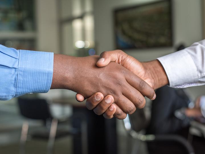 Two people in dress shirts shake hands in front of an office desk.