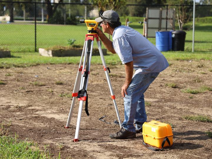 Man in t-shirt and denim jeans crouches to look through a contractor's survey tool.