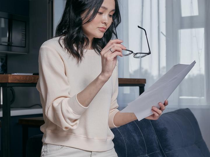 Woman in tan outfit looking through glasses at papers in her hand.