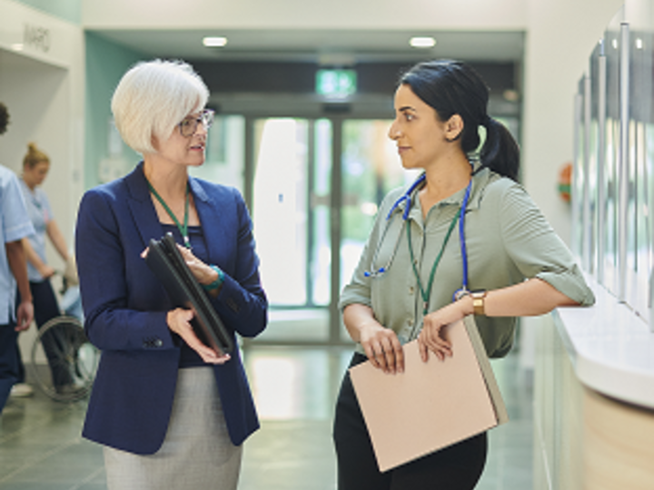 Health care administrator with clipboard talking to medical professional near nurses station.