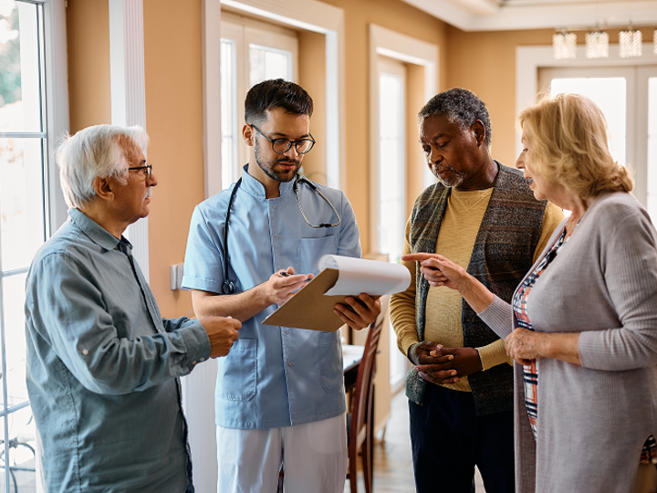 Male nurse reading medical paperwork with group of seniors in residential care home.