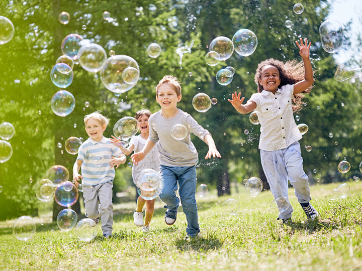Multi-ethnic group of friendly children with toothy smiles on their faces enjoying warm sunny day while playing in soap bubbles.