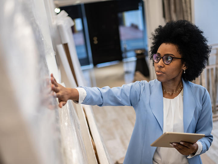 An African American woman in a white shirt and blue blazer touches a white board with her right hand as she holds a tablet in her left hand and gives a presentation.
