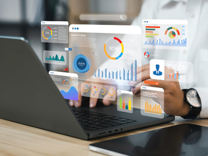 A person in a white button down shirt is typing on a laptop with a tablet sitting on the table next to the laptop. There are graphs and pie charts floating above the keyboard.