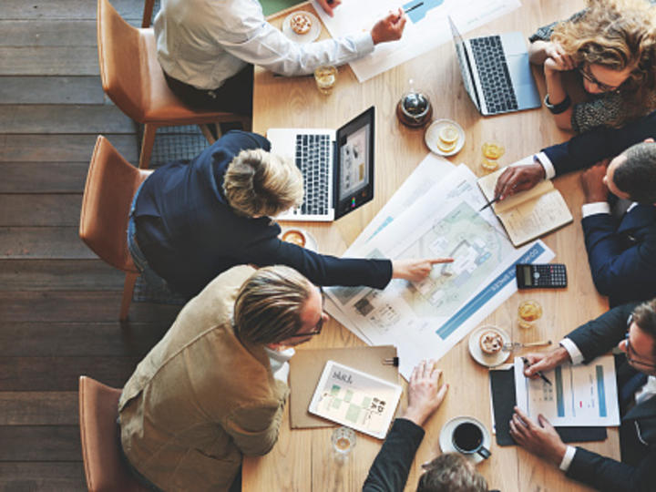 A group of young business professionals are sitting at a conference table with papers, laptops, and coffee cups. The camera is angled from above. A few people are pointing at a large paper in the center.