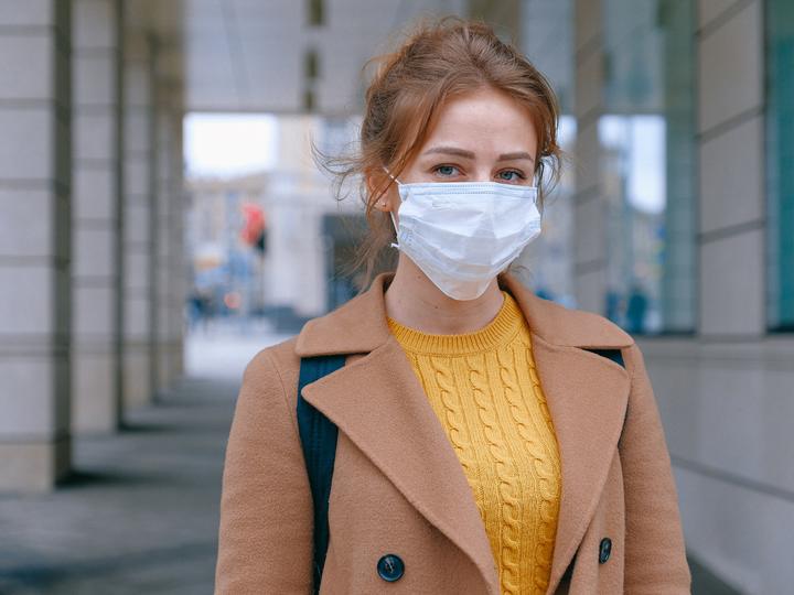 Image of a young caucasian woman wearing a mask and standing outside a building.