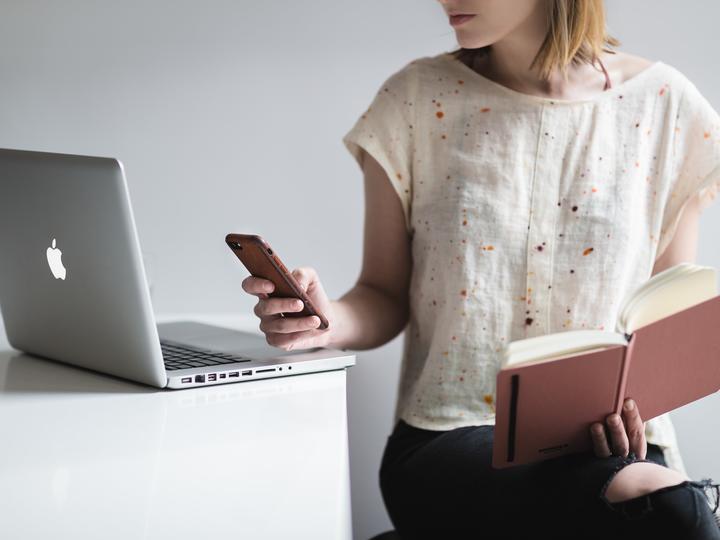 Closeup image of a young caucasian woman checking her phone with her laptop open on a table beside her and an address book in her other hand.
