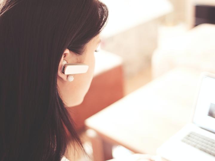 Over-the-shoulder image of a young caucasian woman working on a laptop with a bluetooth earpiece in her ear.