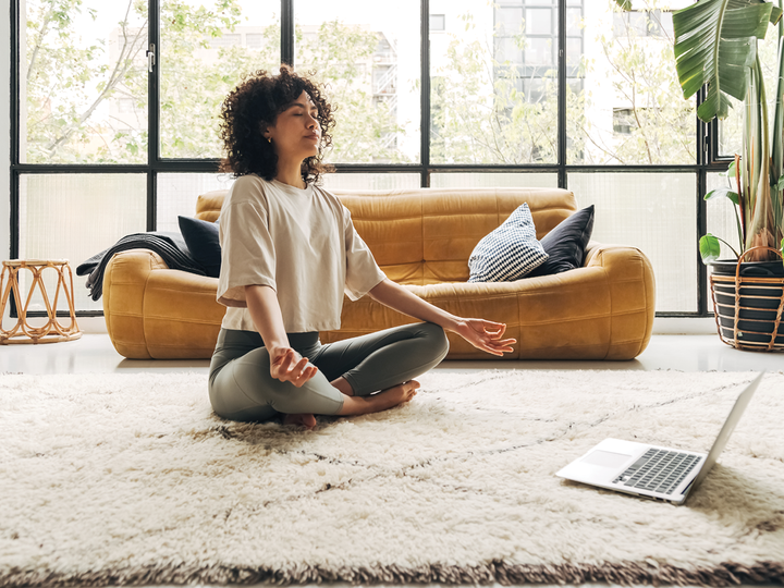 A young woman meditates in lotus pose on the floor of her apartment. Her laptop is open in front of her.