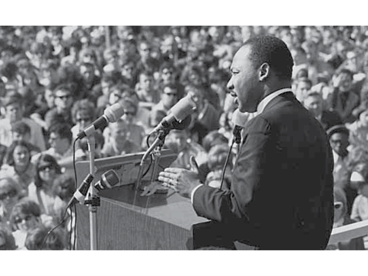 Black and white image of Martin Luther King Jr. at a podium giving a speech with a crowd of blurry faces in the background.