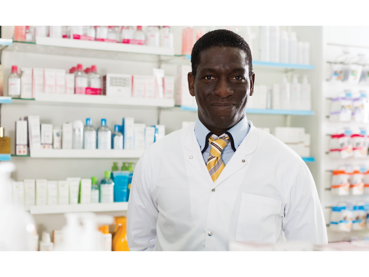 Waist-up image of a male African American pharmacist smiling as he stands in front of the medicine shelves and behind the checkout counter.