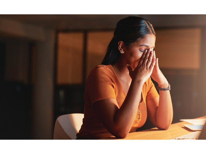 A middle aged woman holds her face in her hands as she sits in front of a lit computer screen in an otherwise dark room.