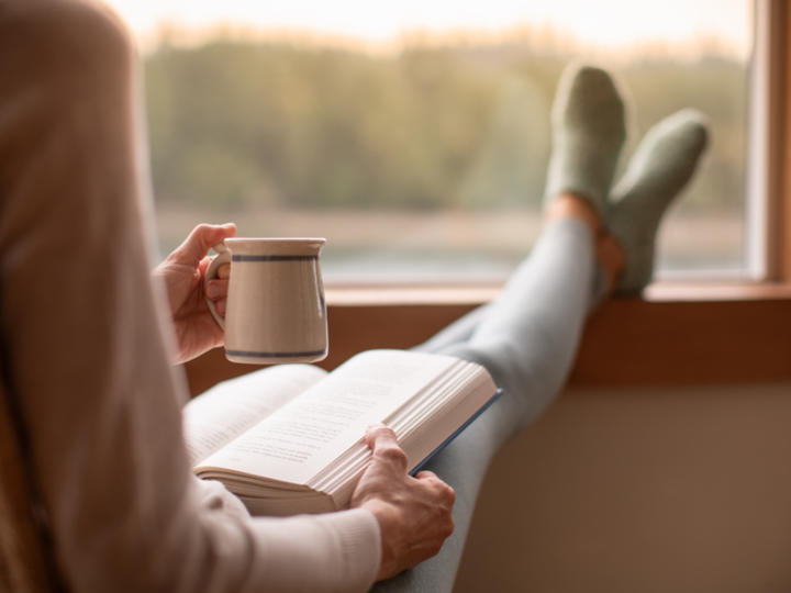 Photo of person from the back holding a mug and open book facing a window with legs propped up