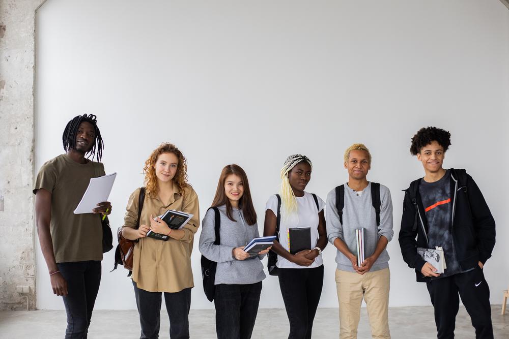 Six diverse young people wearing bookbags standing shoulder to shoulder. Each person is holding at least one notebook. Their faces are warm and smiling.