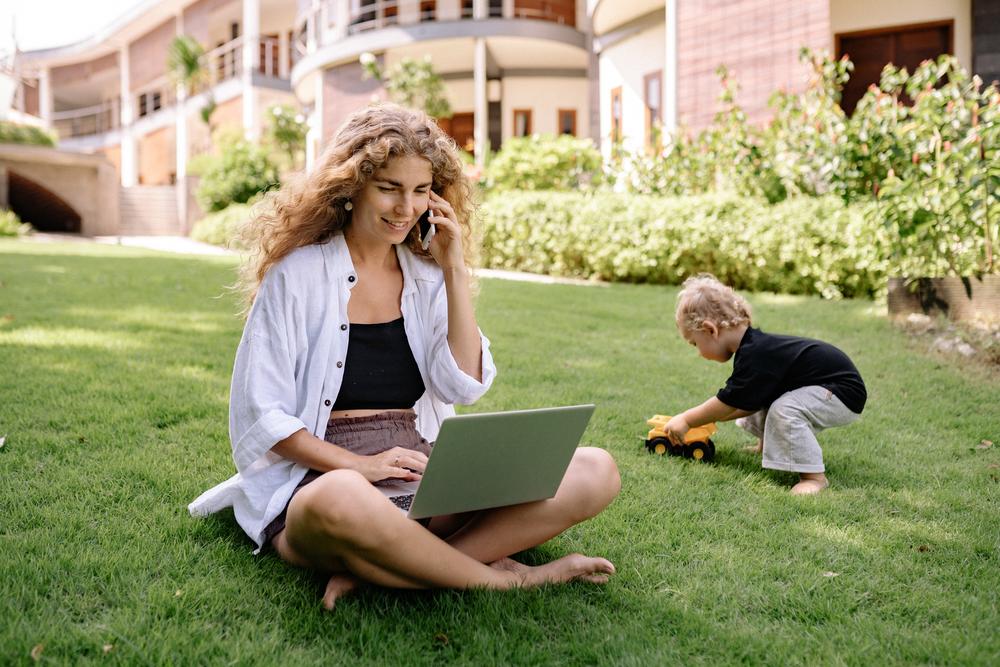 Barefoot woman sitting on grass outside large home on a sunny day. She smiles while talking on the phone and using a laptop. Her barefoot toddler plays with a toy truck in the background.