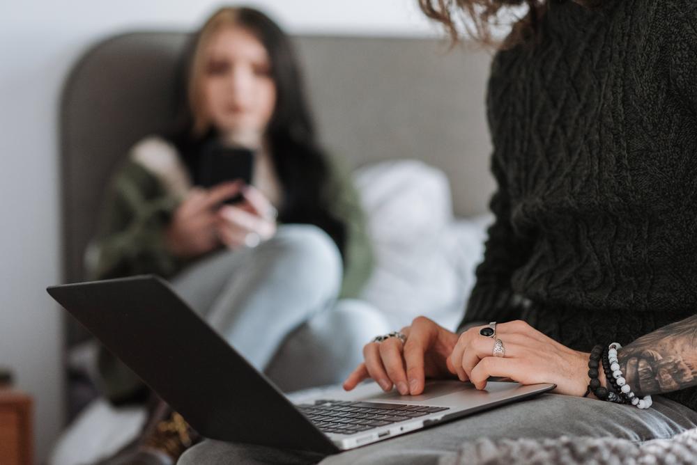 Young couple sitting on bed. In the foreground, man with tattoos uses a laptop. In the background, dark-haired woman uses cellphone.