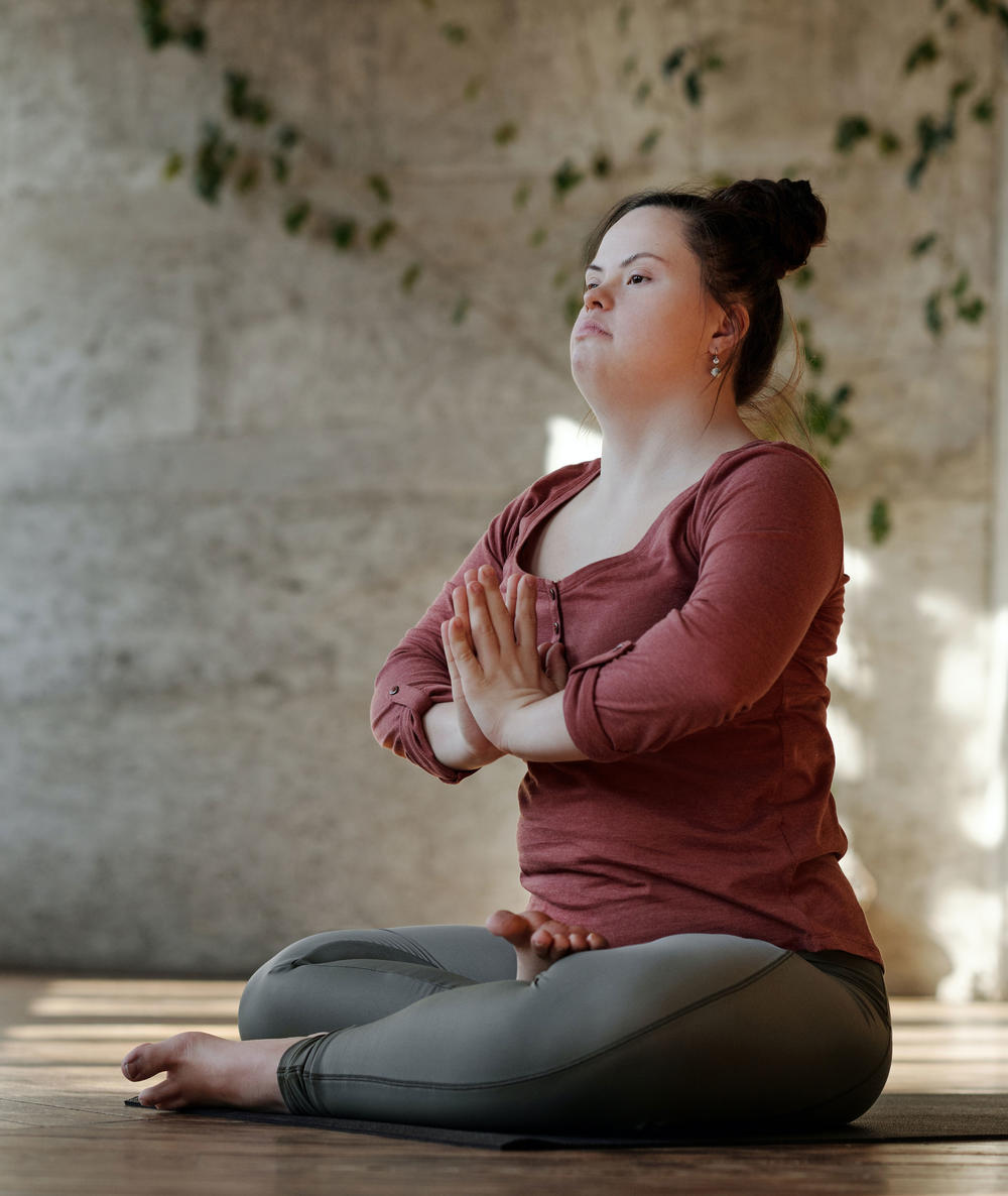 Young woman with down syndrome sitting on the floor with her legs crossed and her palms together, practicing yoga. Her expression is focused and calm.