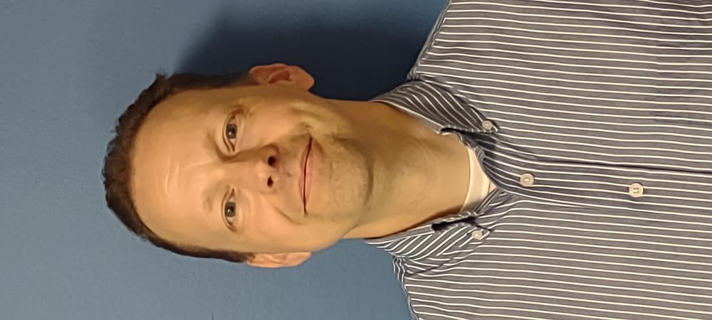 A smiling man in a gray and white striped collar shirt in front of a gray background.