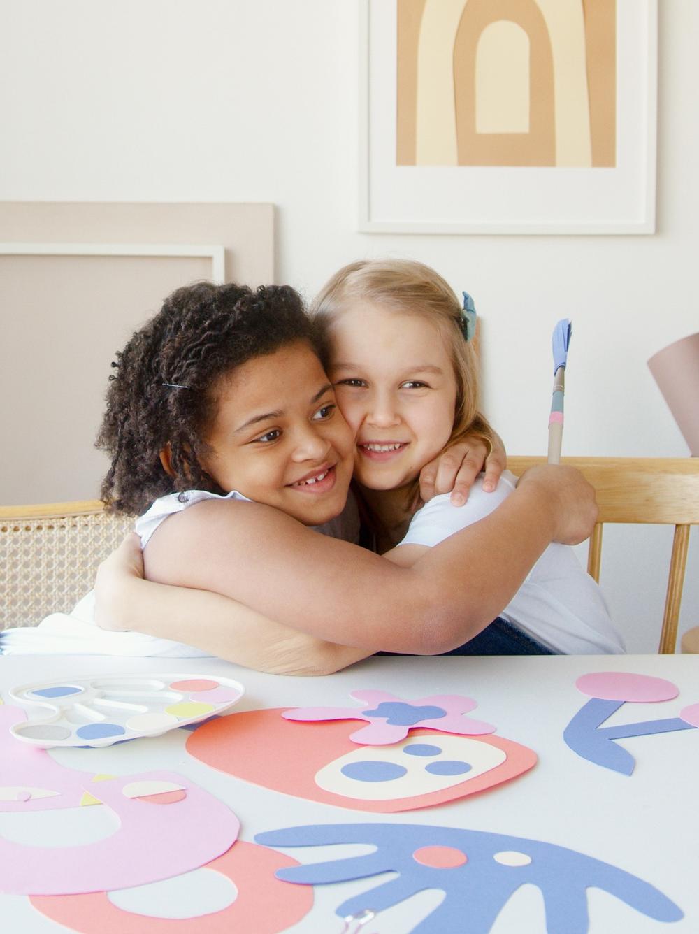Two young girls smiling and hugging at an arts and crafts table.