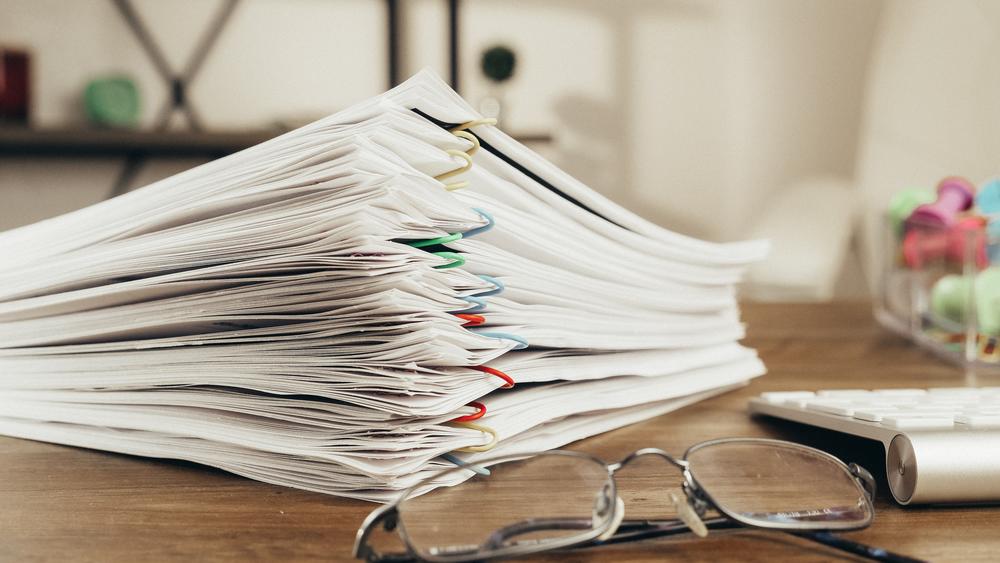 Stacked papers grouped together with multicolored paperclips on a wooden desk next to glasses and a wireless keyboard.