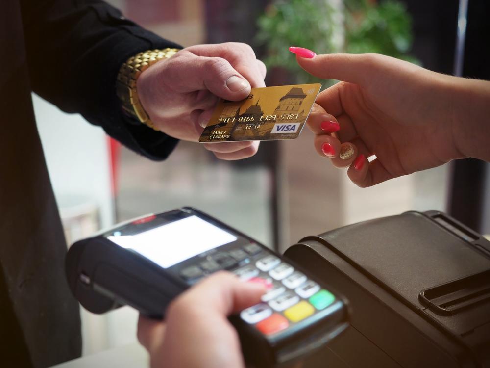 Person in business suit with gold watch handing a gold Visa credit card to a person with red nails holding a card swipe terminal.