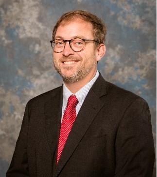 Man smiling at camera wearing glasses and a dark brown suit with a red tie.