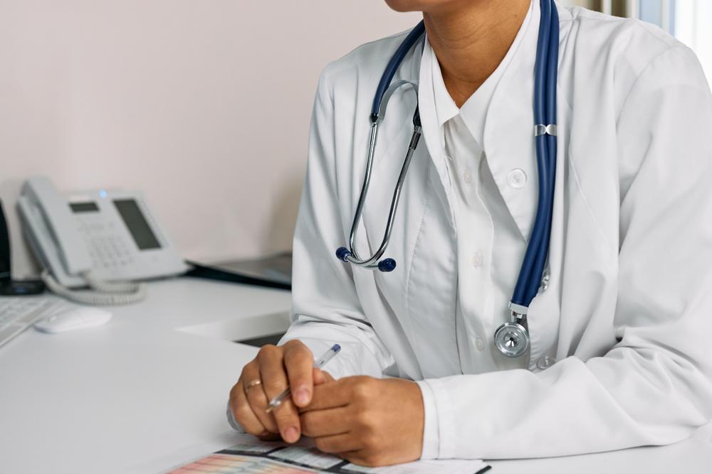 Person wearing stethoscope and doctor's white coat sitting at desk with pen and paper.