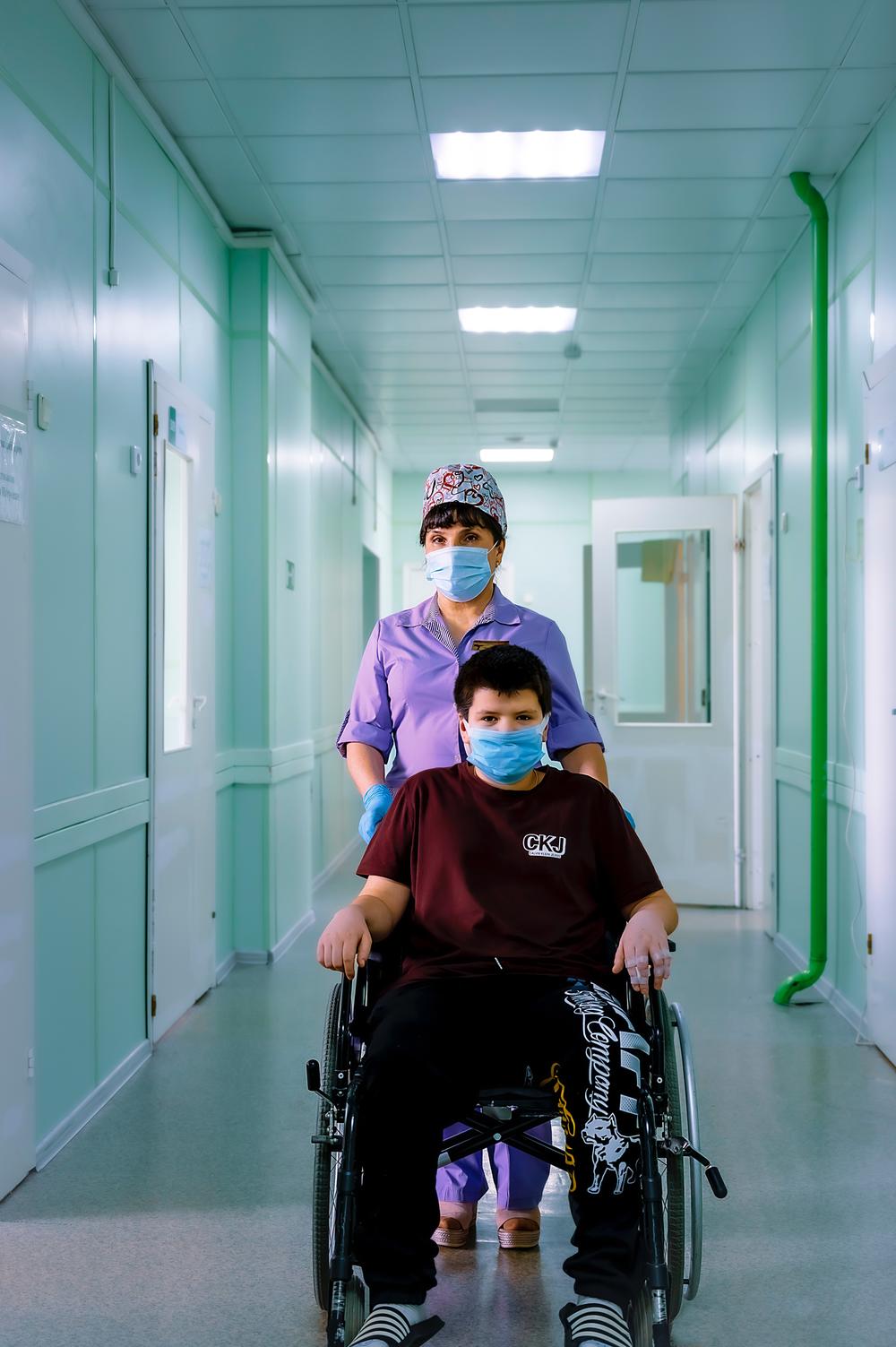 Nurse in purple scrubs and mask walking patient in mask and wheelchair down a facility hallway.