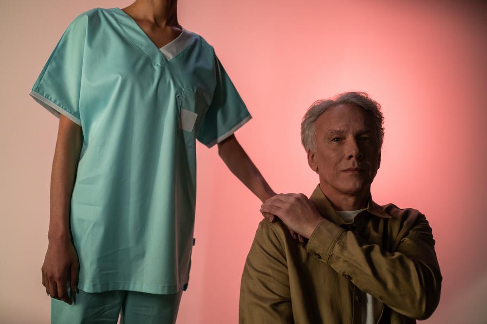 Nurse standing with hand on shoulder of calm elderly man sitting beside them.
