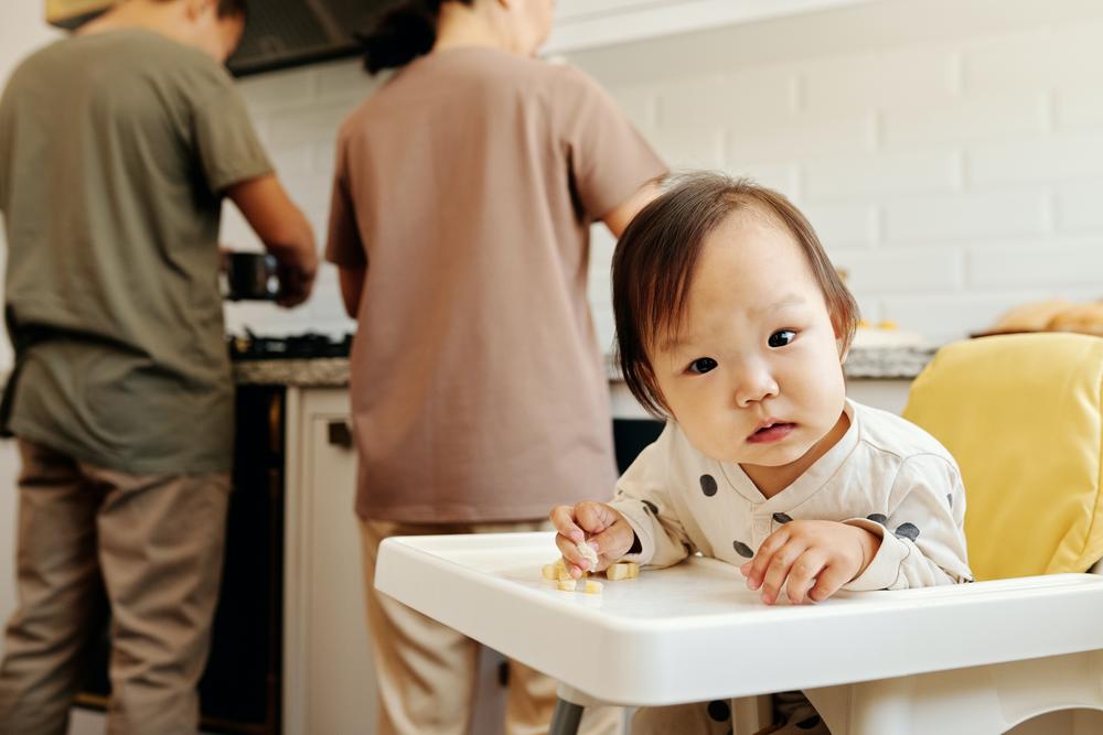 Parents cooking in background while baby looks curiously at camera.