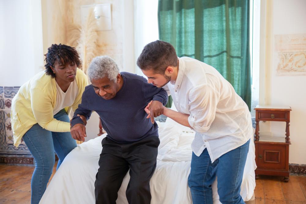 Two nurse aides helping an elderly man stand up at home.