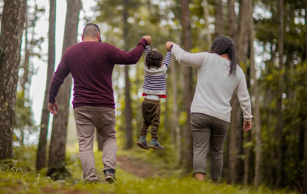 Parents walking in forest with child between them, lifting child by arms.
