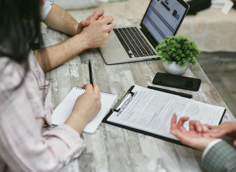 Three people sitting at a table with a computer, notebook and paperwork.