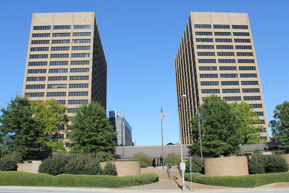 Wide-angle shot of two exactly identical pale brick buildings with small rectangular windows and a plaza with an American flag and trees in front.