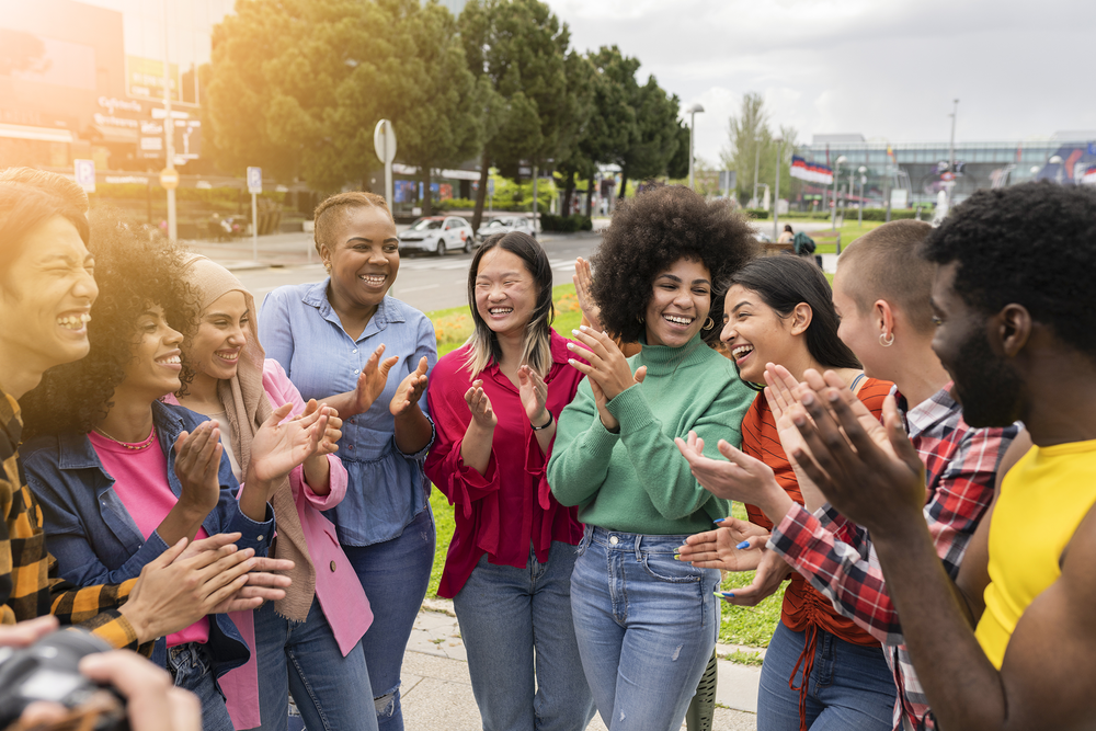 A diverse group of young people stand in a park in a half-circle applauding and smiling at one another.