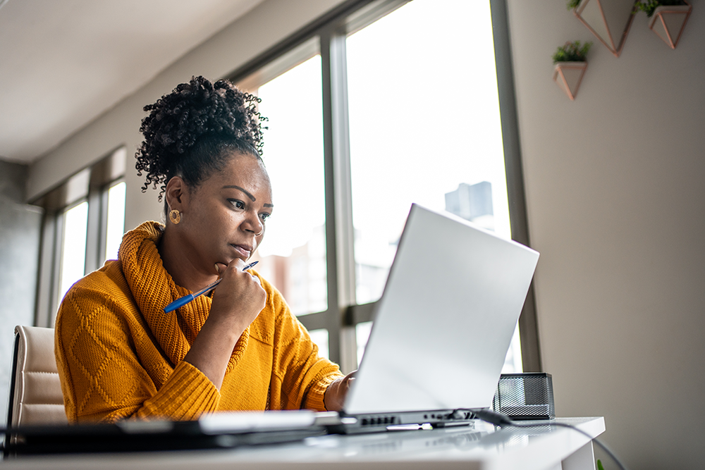 Image of a middle aged African American woman staring intently at her laptop screen. She sits in a brightly lit apartment and has a pen in her hand for taking notes.