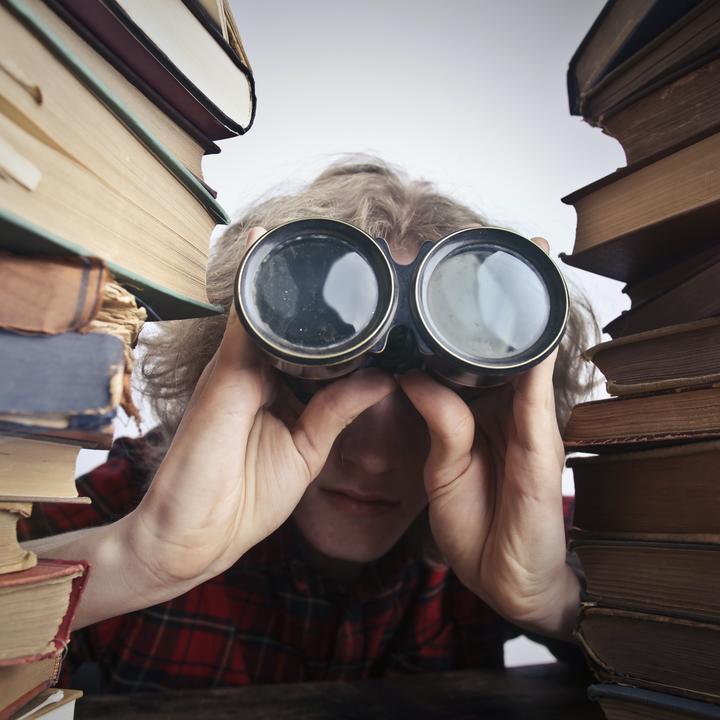 A person looking through binoculars between two towering stacks of old books.