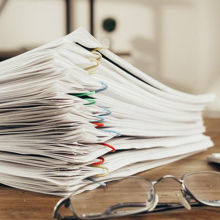 Stacked papers grouped together with multicolored paperclips on a wooden desk next to glasses and a wireless keyboard.