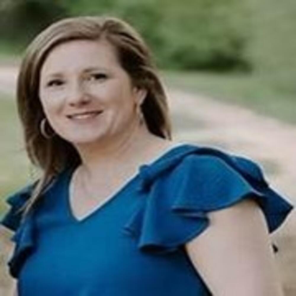 Photo of woman wearing a blue shirt, smiling out side with greenery in the background.