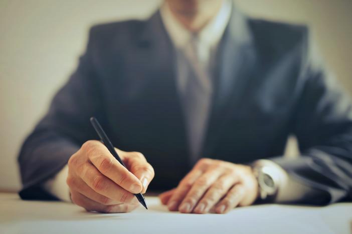 Close up of man's hand holding pen as if to write. Blurred background shows man is wearing suit.